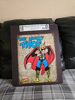 Front view of the metal sign showing a colorful comic style image of The Mighty Thor with a rustic look background and attached to black cardboard backing with label.