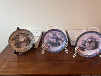 Three collector plates on stands with their boxes in background shown on table.