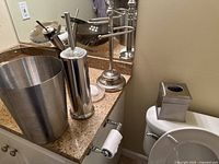 Photo showing stainless steel waste basket, toilet brush holder, towel rack, tissue holder on and near bathroom counter and toilet.