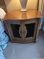 Front view of one wooden bedside table with cabinet doors showing metal mesh inserts and decorative handles, standing on carpet next to bed.