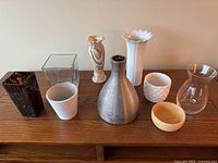Photo showing 9 vases and bowls of different styles and materials arranged on wooden surface against beige wall.