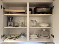 Open kitchen cabinet showing three shelves with assorted kitchenware: cutting boards, baking trays, kitchen utensils, ceramic plates, bowls, glass jar and measuring cup, mortar and pestle, vegetable chopper in box.