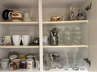 Wide view of upper shelf showing ceramic mugs, a ceramic pitcher, decorative vases, and multiple clear glass cups, including dessert or cocktail glasses.
