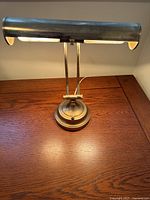 Photo of one brass desk lamp on a wooden surface, showing the cylindrical lamp head and light illuminated.