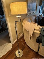 Full view of the floor lamp standing next to couch with beige fabric shade and chrome finish.