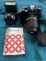 Photo showing Nikon EM camera with zoom lens mounted, Vivitar 283 flash, and Vivitar user manual on blue cloth background.