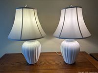 Two matching white ceramic table lamps with ribbed vase-shaped bases and white fabric bell-shaped shades sitting on a wooden table.