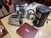 Various kitchen appliances on countertop including chrome toaster, stainless steel kettle, black coffee maker, Braun Multimix boxed, and wooden wine accessory box.