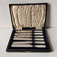 Set of six sterling silver butter knives with mother of pearl handles arranged in original blue velvet-lined box with cream satin lid lining.