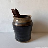 Front view of stoneware utensil jug with dark tenmoku glaze and some wooden utensils inside for scale.