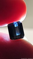 Close-up image of the blue-purple emerald-cut tourmaline held between finger and thumb showing its size and color under natural light.