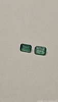 Pair of octagon cut green emerald gemstones on a neutral surface under natural light showing their clarity and color.