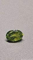 Oval cut natural green peridot gemstone approx 3.38 carat lying on textured background showing apple green color with inclusions
