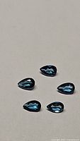Five deep blue pear-shaped London blue topaz gemstones arranged on a neutral background, displaying their shape, size, and color.