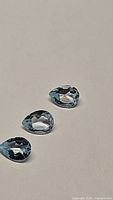 Three pear-shaped sky blue topaz gemstones arranged in a line on a plain beige surface.