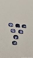Seven calibrated oval cut tanzanite gemstones laid out on a flat surface, showing their blue to purple color and transparency under direct light.