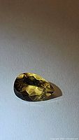 Close-up photo of a pear-shaped yellow Helidor gemstone on a plain surface showing its facets and golden yellow color under diffused lighting.