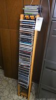 Wooden vertical CD tower fully loaded with music CDs, standing against a wooden wall and near a filing cabinet.