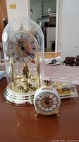 Birks anniversary clock under glass dome with small German alarm clock and music box piano in front on table