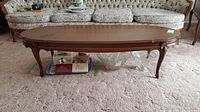 Front side view of the oval wooden coffee table in a room with floral couch and carpet. Surface shows scratches.