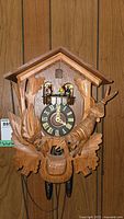 Front view showing entire cuckoo clock mounted on wood paneling, featuring carved bird, large leaves, black clock face with gold Roman numerals, and decorative dancing figurines balcony.