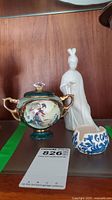 Porcelain container with lid and gold-tone handles, white Japanese figurine, and small blue-white dip dish on wooden shelf