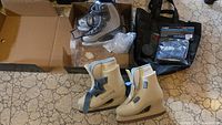 Photo showing two pairs of ladies ice skates (Reebok size 5 and Lange size 5-6) with some packaging and accessories on a tiled floor.