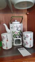 Full tea set containing teapot with brass handle, ceramic warmer stand, and four matching cups with decorated floral and calligraphy designs.