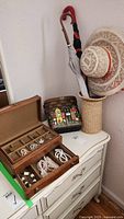 Photo showing wooden jewelry box open with white bead jewelry inside, butterfly pin visible, storage basket holding umbrellas and hats nearby