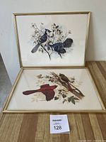 Two framed bird prints displayed on a wooden table, one featuring blue jays, the other red cardinals with butterflies and floral background.