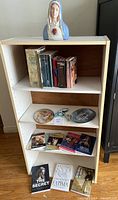 Full bookshelf showing four shelves with books, religious plates, rosary, and top Virgin Mary statue