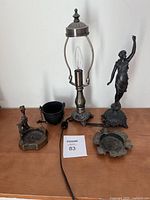 Full view of all items: shadeless lamp, cast iron pot, bronze figurine, and three ashtrays on wood surface.