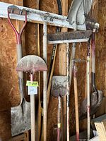 Photo of 9 assorted garden tools hung on a white rail inside a wooden shed, showing shovels, rakes, and a broom with visible dirt and signs of use.