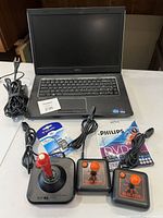 Dell laptop open with keyboard, power cord, three joysticks, Verbatim SD card, and Philips DVD-R discs on table.