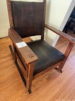Front view of vintage wooden rocking chair with black leather seat and backrest showing wear and a removable seat cushion.