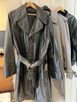 Three men's coats hanging on wall hooks, front view showing black leather trench coat in front, gray trench coat and black leather jacket behind it.