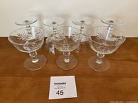 Seven clear crystal stem glasses with pin wheel cut pattern on bowl showing starburst design, arranged in two rows, with one glass possibly chipped on the rim.