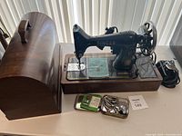 Full setup of Singer 99K sewing machine on wooden base with power cord and various metal accessory tins, alongside curved wooden Singer case.