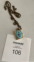 Full length view of the book locket pendant with floral blue medallion on chain with MaxSold lot card.