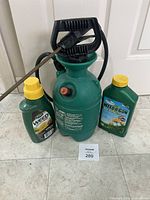 Green plastic 1-gallon Garden Club sprayer with pump handle, spray wand, and two herbicide bottles placed next to it.