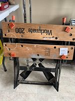 Front view of the Black & Decker Workmate 200 foldable workbench with wooden top and black steel frame, showing wear and orange plastic clamps.