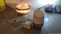Google Home smart speaker on a table with integrated power cable and connected power adapter with 'G' logo.