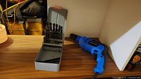 Photo of blue Mastercraft corded drill and metal tin with drill bits on a wooden surface under overhead lighting.