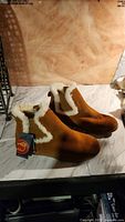 Pair of tan suede men's ankle boots with white shearling lining around the ankle. Rubber sole and pull tab at heel visible. Lamo Footwear tag attached.