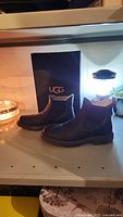 Side and angled view of men's dark brown UGG boots showing elastic panels and sole tread with UGG box behind.