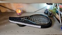 Prince tennis racket placed on top of its black zippered case on a flat surface with a lit candle and plants in the background.