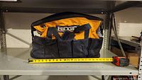 Front view of the tool bag showing RIDGID logo, multiple front pockets, and a tape measure placed below for width reference showing approx 22 inches