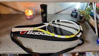 Babolat tennis racket resting on Babolat carrying bag with Wilson tennis ball can.