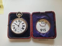 Pocket watch displayed in open case with blue velvet lining and a removable circular tag showing lot number.
