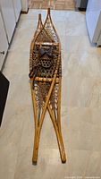 Pair of antique wooden snowshoes placed on a light floor. Visible wood frame, rawhide netting, and leather bindings.